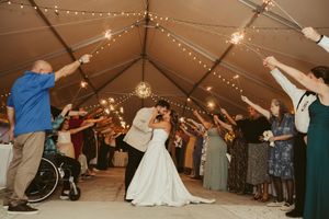 Bride and groom share a romantic dip kiss during sparkler exit at wedding reception under a tent with string lights.