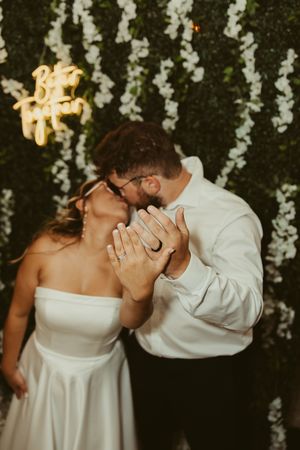 Newlyweds kiss and display wedding rings in front of a floral green wall with a glowing neon sign.