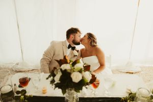 Bride and groom share a kiss at sweetheart table with floral centerpiece at reception.
