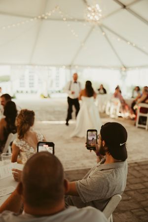 Wedding guest photographs father-daughter dance on smartphone at tented reception celebration.