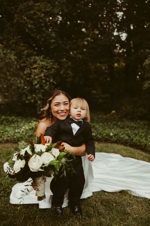 Smiling bride in strapless gown holds white bouquet while posing with toddler in tiny tuxedo outdoors.