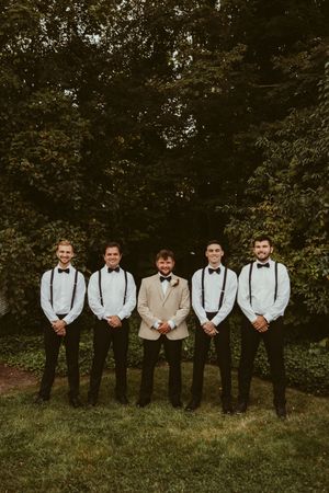 Groom in tan blazer poses with four groomsmen in white shirts and suspenders in a garden setting.