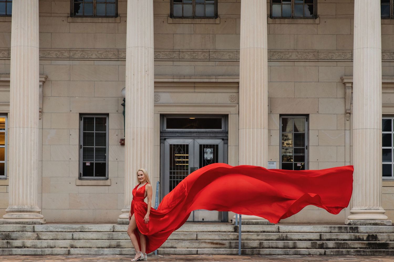 A flowing red dress billows dramatically against a pink building with palm trees in a tropical setting.