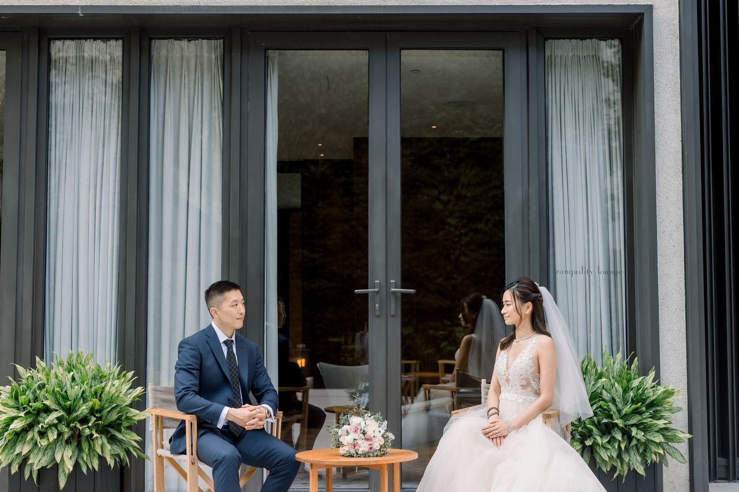 A bride and groom share a romantic moment outside a modern building with large windows and potted plants.