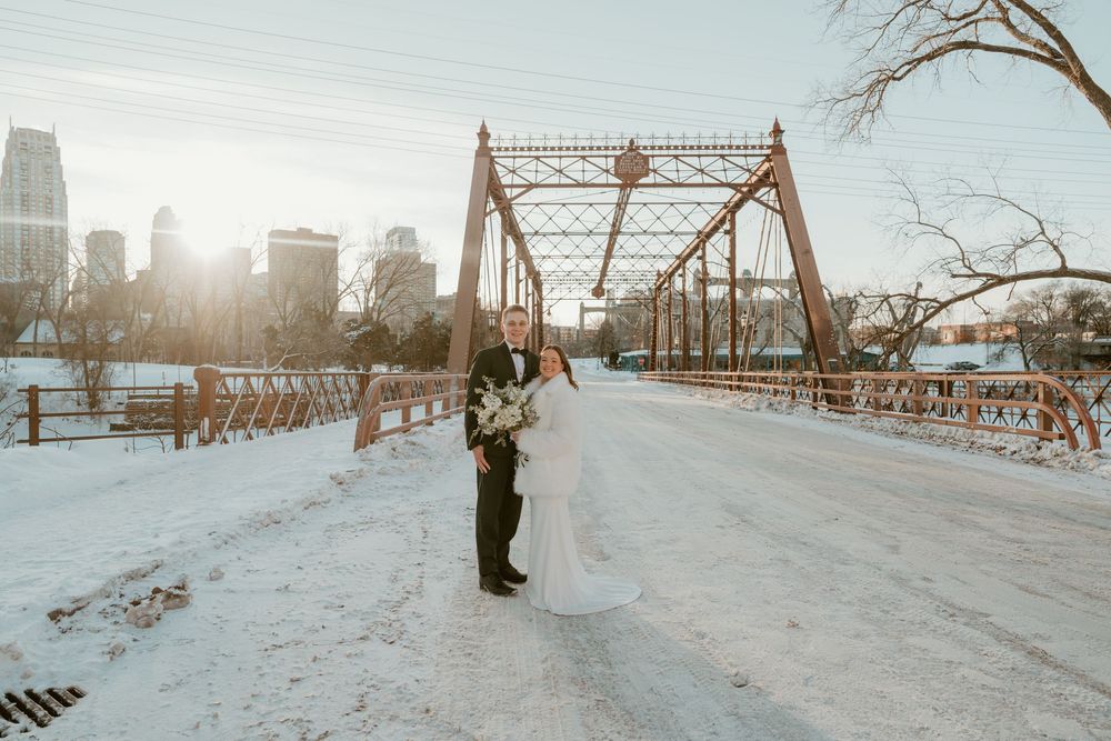Winter wedding couple stands in snowy park with urban skyline and bridge in background at sunset.