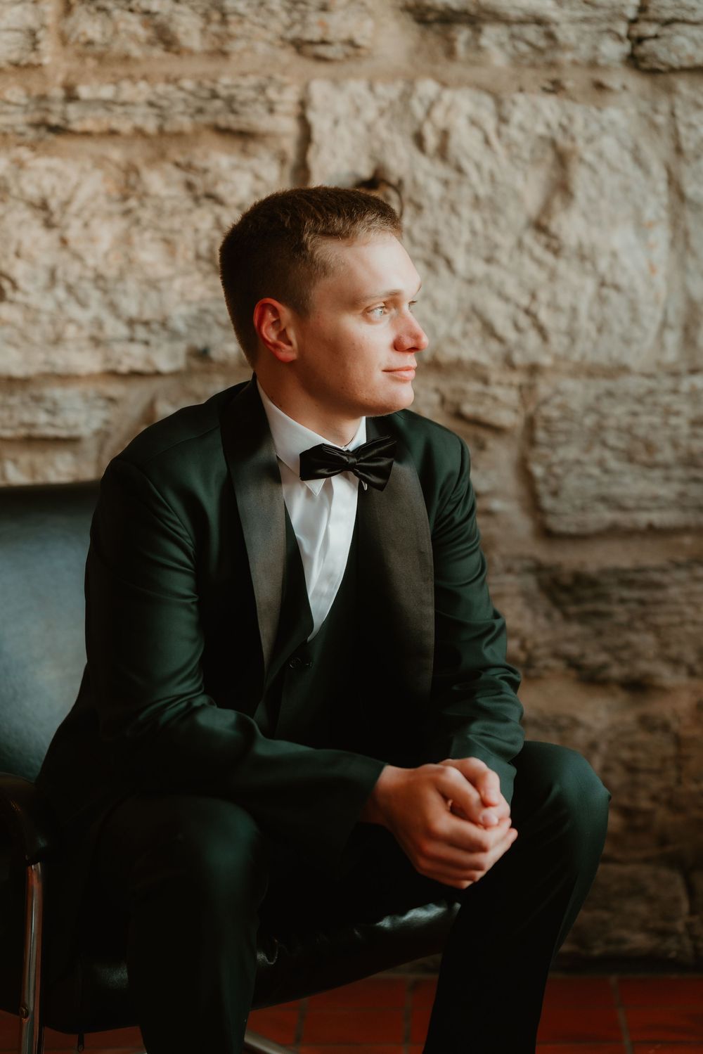 A person in an elegant dark green velvet tuxedo and bow tie sits thoughtfully against a stone wall background.