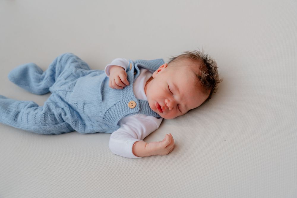 A sleeping baby wearing a light blue outfit lies on a soft white surface in a peaceful pose.