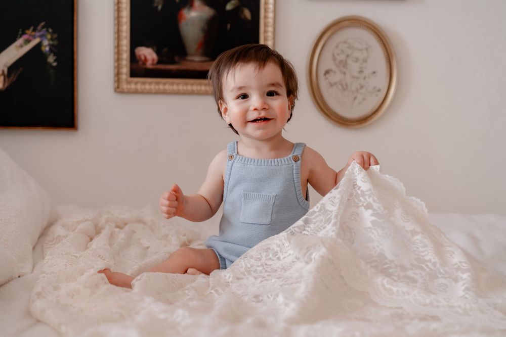 A baby in a light blue outfit plays with flowing white tulle fabric while sitting on a bed.