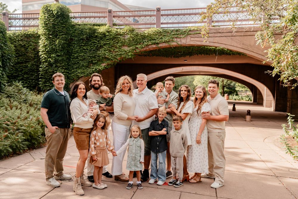 A group of people in light colored outfits pose together under an arched bridge in a garden setting.