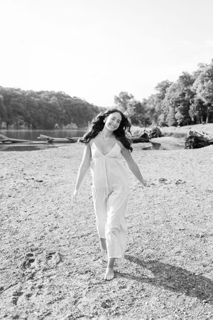 A person in a flowing white dress walks gracefully across a rocky beach landscape in black and white photography.