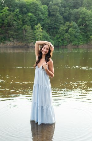A woman wearing a light blue tiered dress stands in shallow lake water surrounded by green trees on the shore.