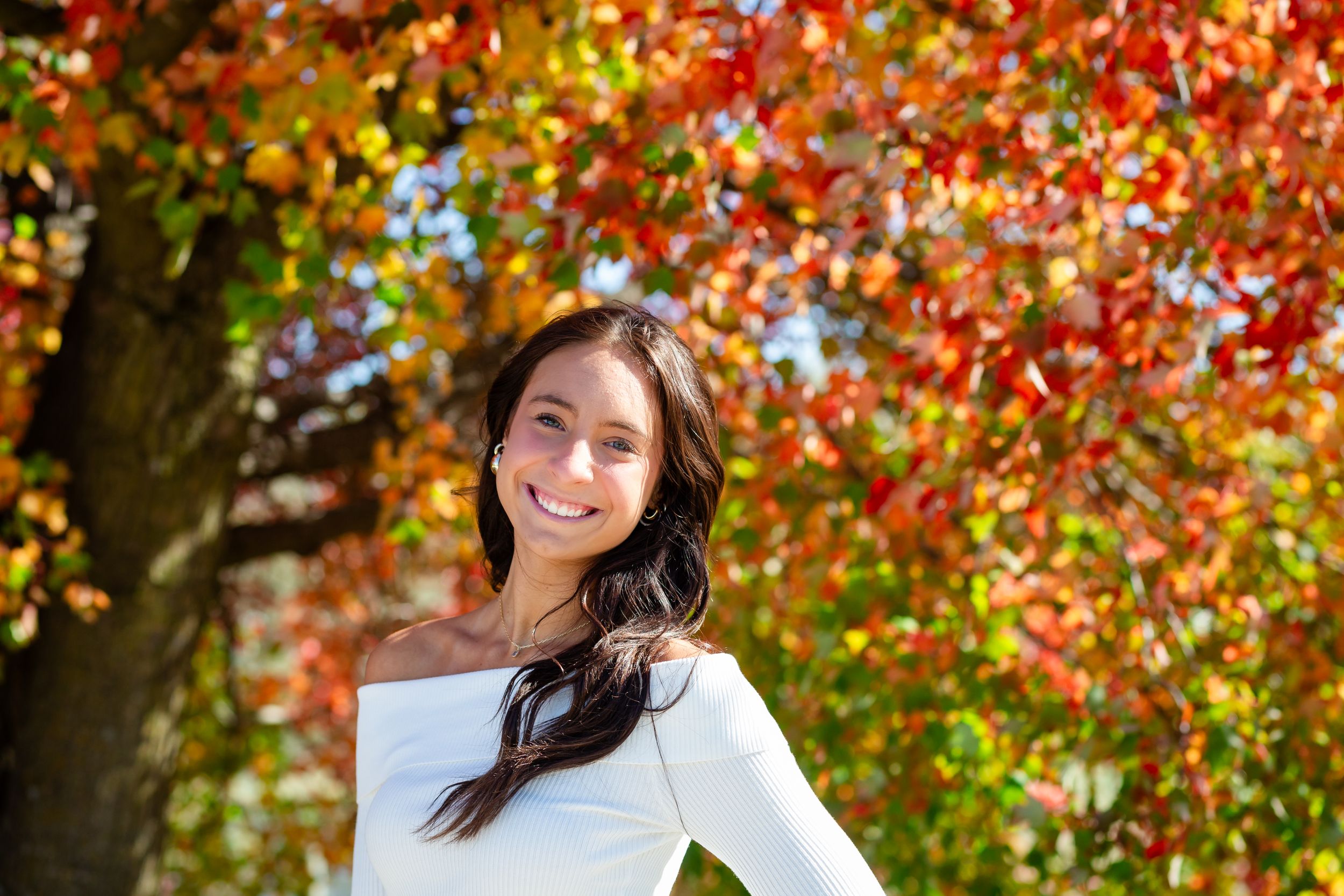 Someone posing outdoors with vibrant red and orange autumn foliage in the background during fall season.