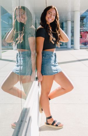 Woman in denim crop top and white skirt poses confidently in bright urban setting with reflective surfaces.