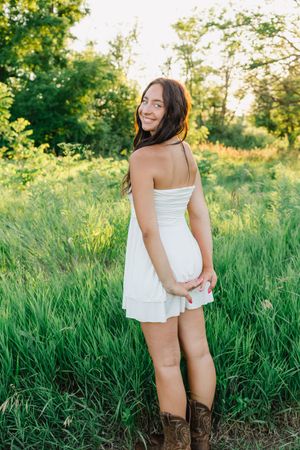 A person in a white sundress and brown boots poses in a sunlit meadow with green grass and trees in the background.