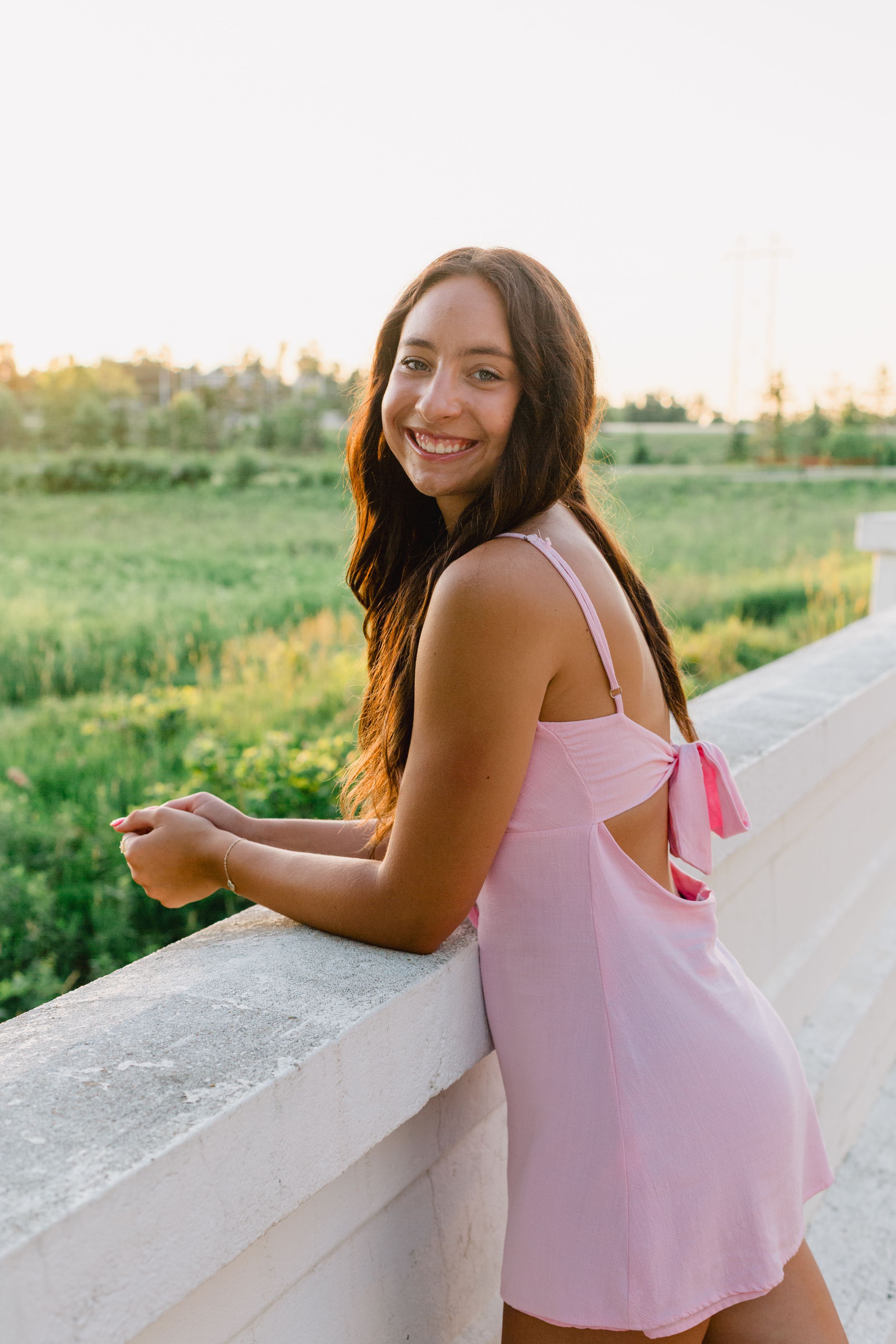 A person in a pink tank top leans against a concrete barrier at sunset with green fields in the background.