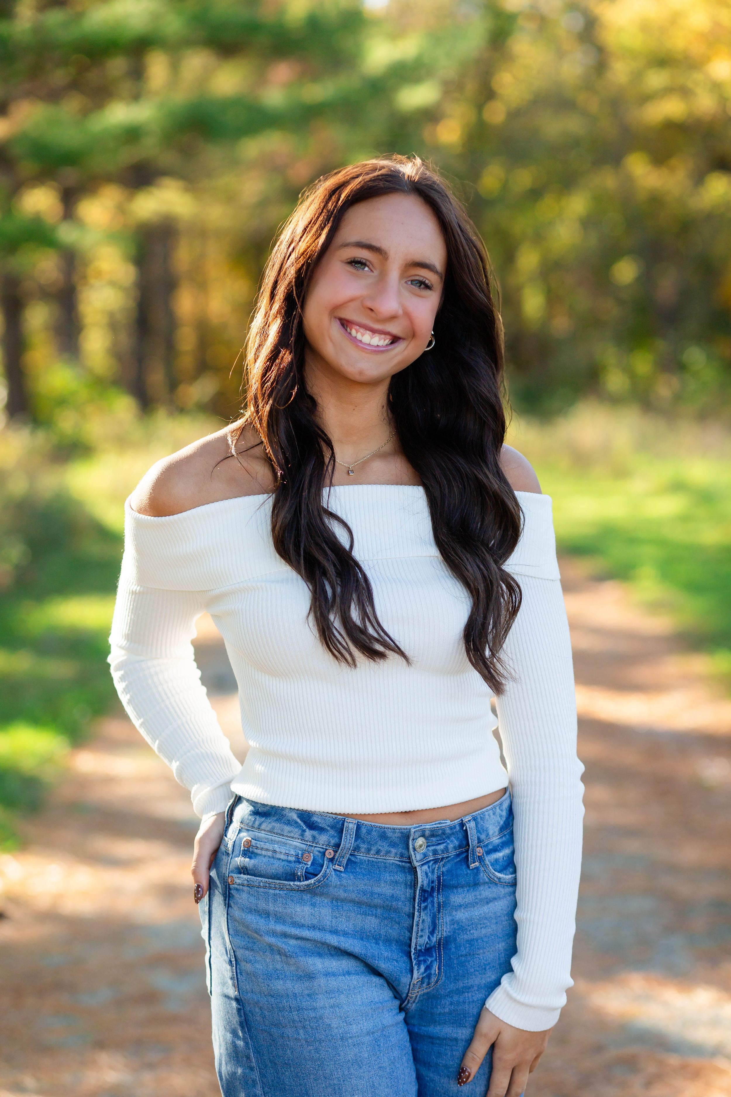 A person wearing a white off-shoulder top and blue jeans poses outdoors in autumn with golden foliage in the background.