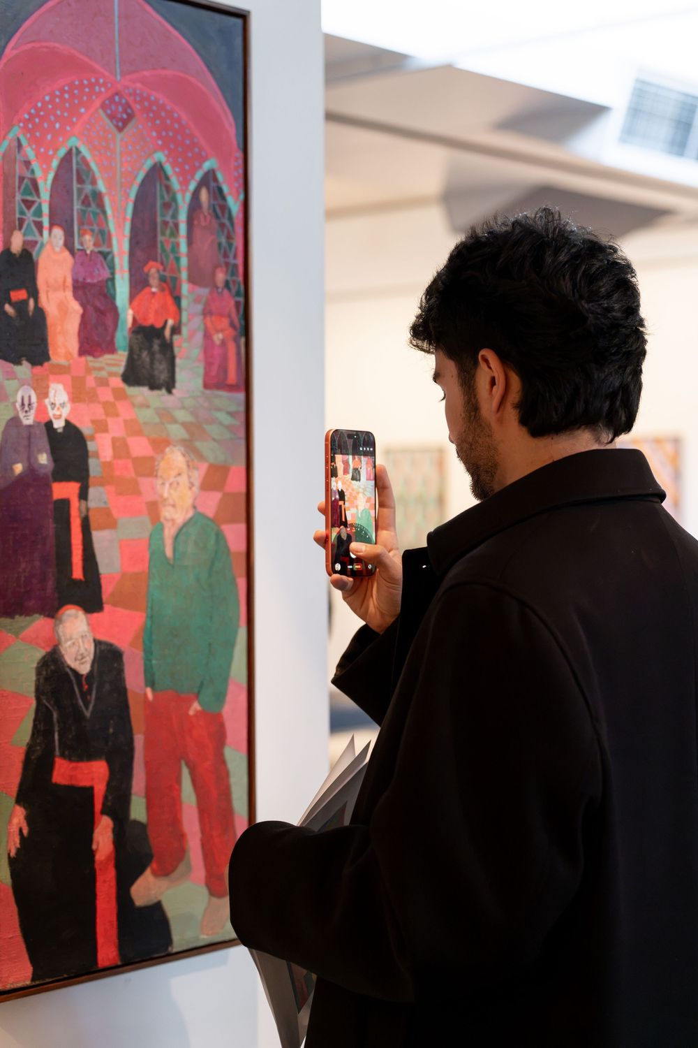 Man in black jacket photographing a colorful figurative painting at an art gallery exhibition.