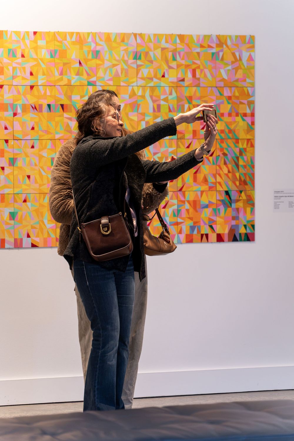 Woman taking a selfie in front of colorful geometric abstract artwork on white gallery wall.