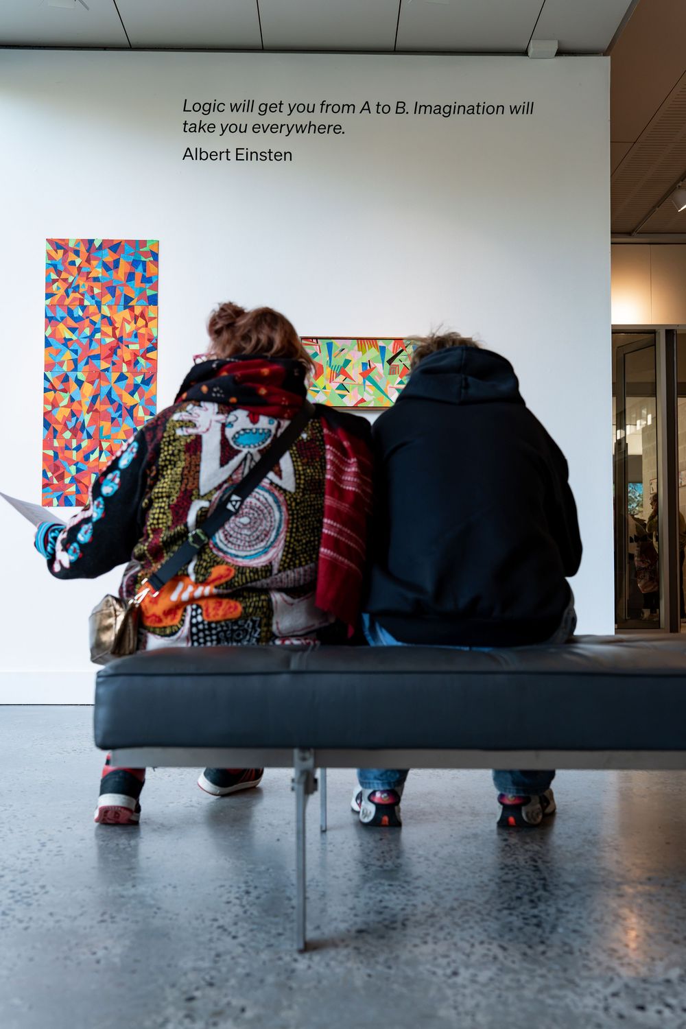 Two museum visitors sit on a bench viewing colorful abstract art with an Albert Einstein quote on the wall.