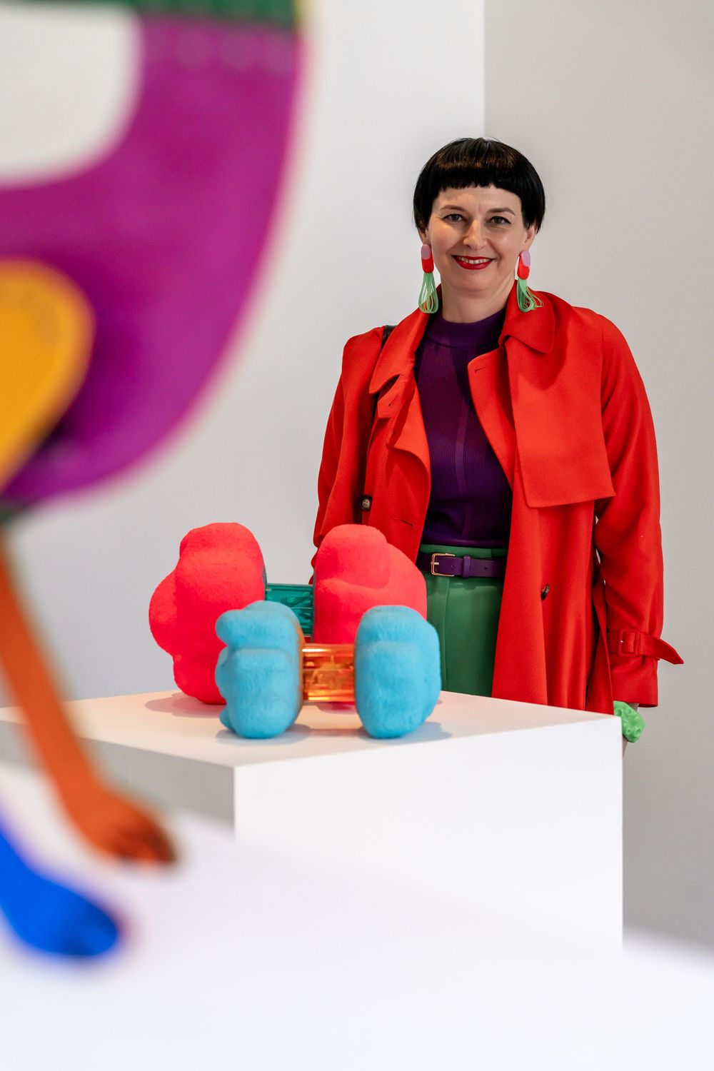 A person in a bright red blazer and purple top stands next to colorful blue objects on a white surface.
