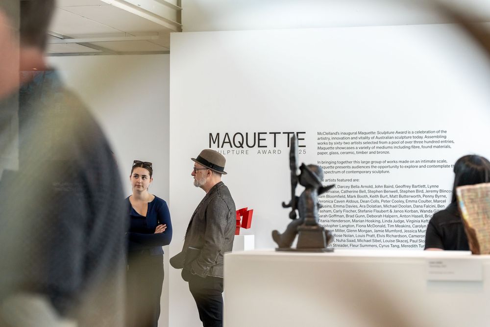 Art gallery exhibition space showing maquette display with visitors viewing wall text and artwork.