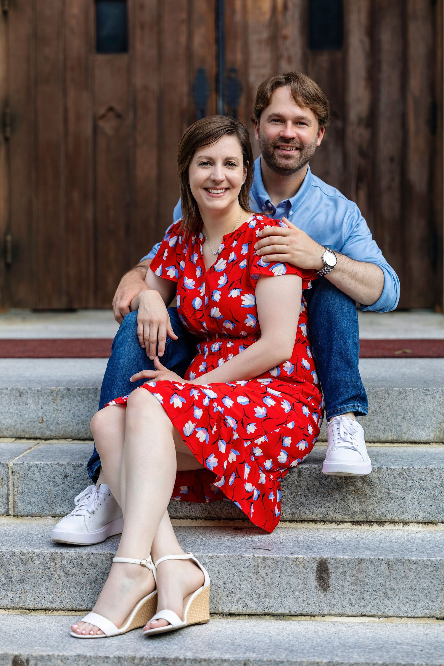 A couple sits together on concrete steps, with her wearing a red polka dot dress and him in a blue button-down shirt.