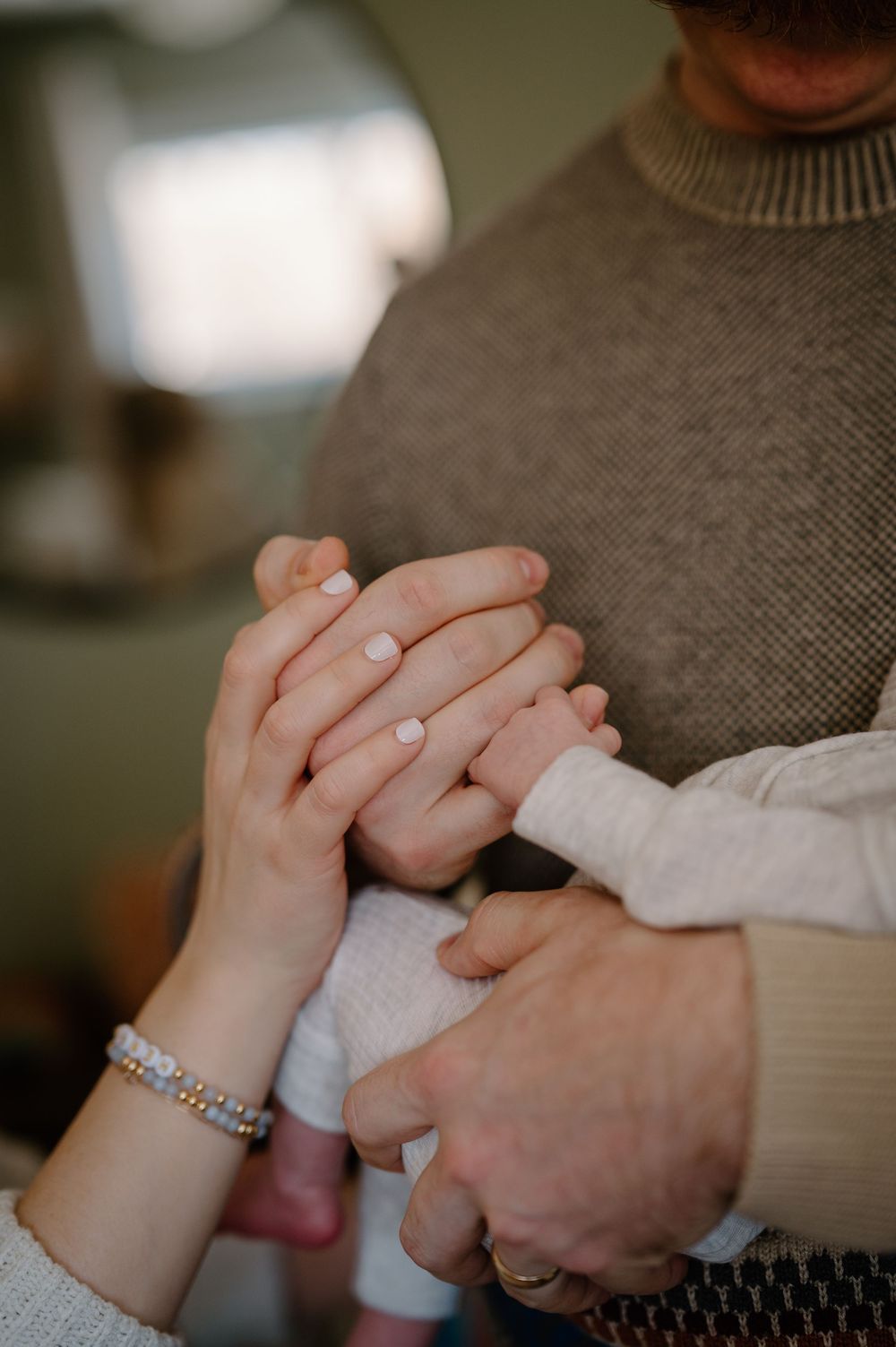 Close-up of hands holding a baby's hand, showing a bracelet and sweater sleeve in soft natural lighting.