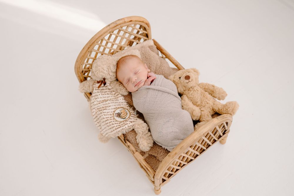 Sleeping baby in cozy wicker bassinet with plush teddy bear wearing neutral colored swaddle.