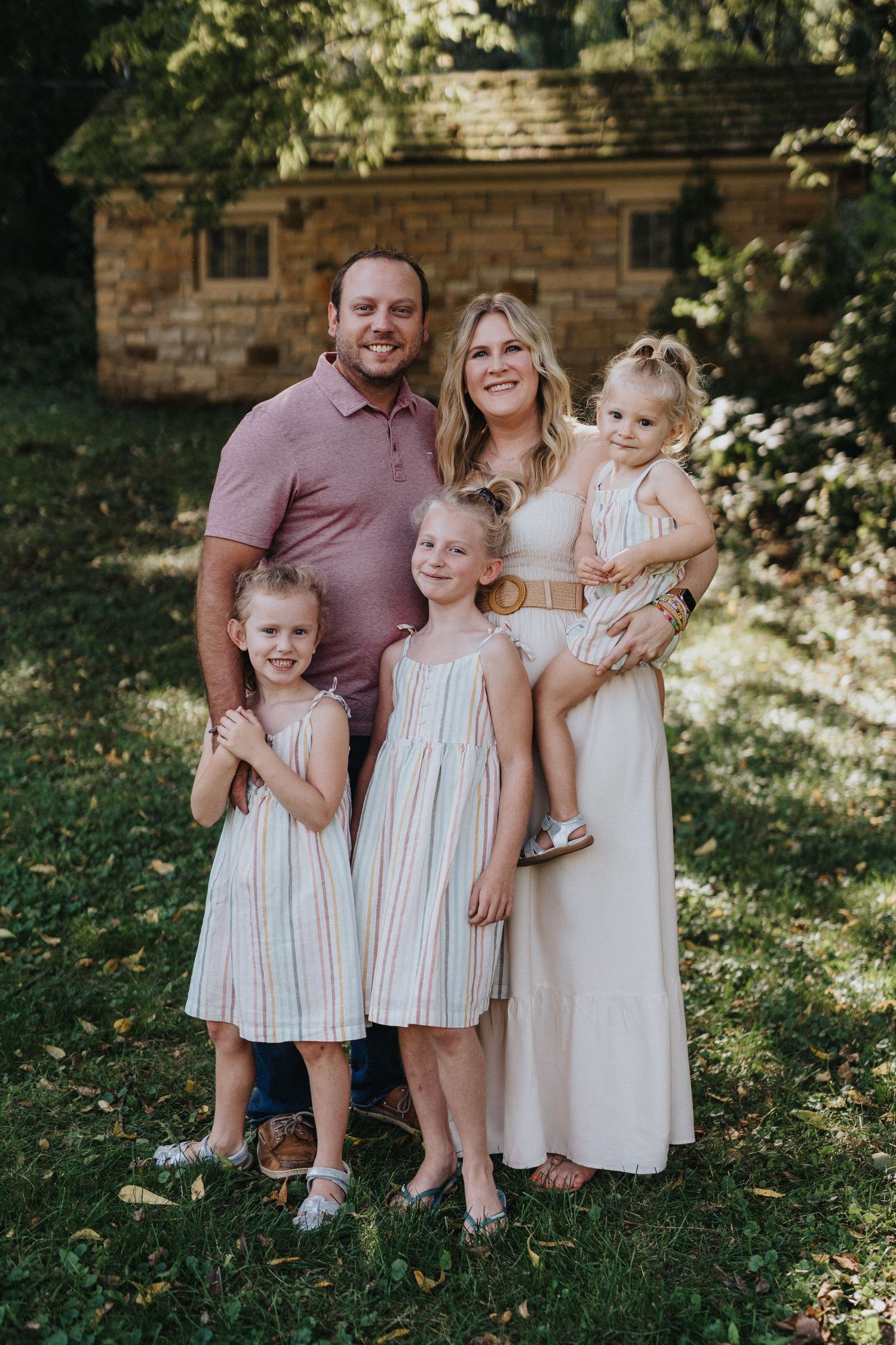 A young family poses together outdoors on a sunny day in front of a rustic stone building.
