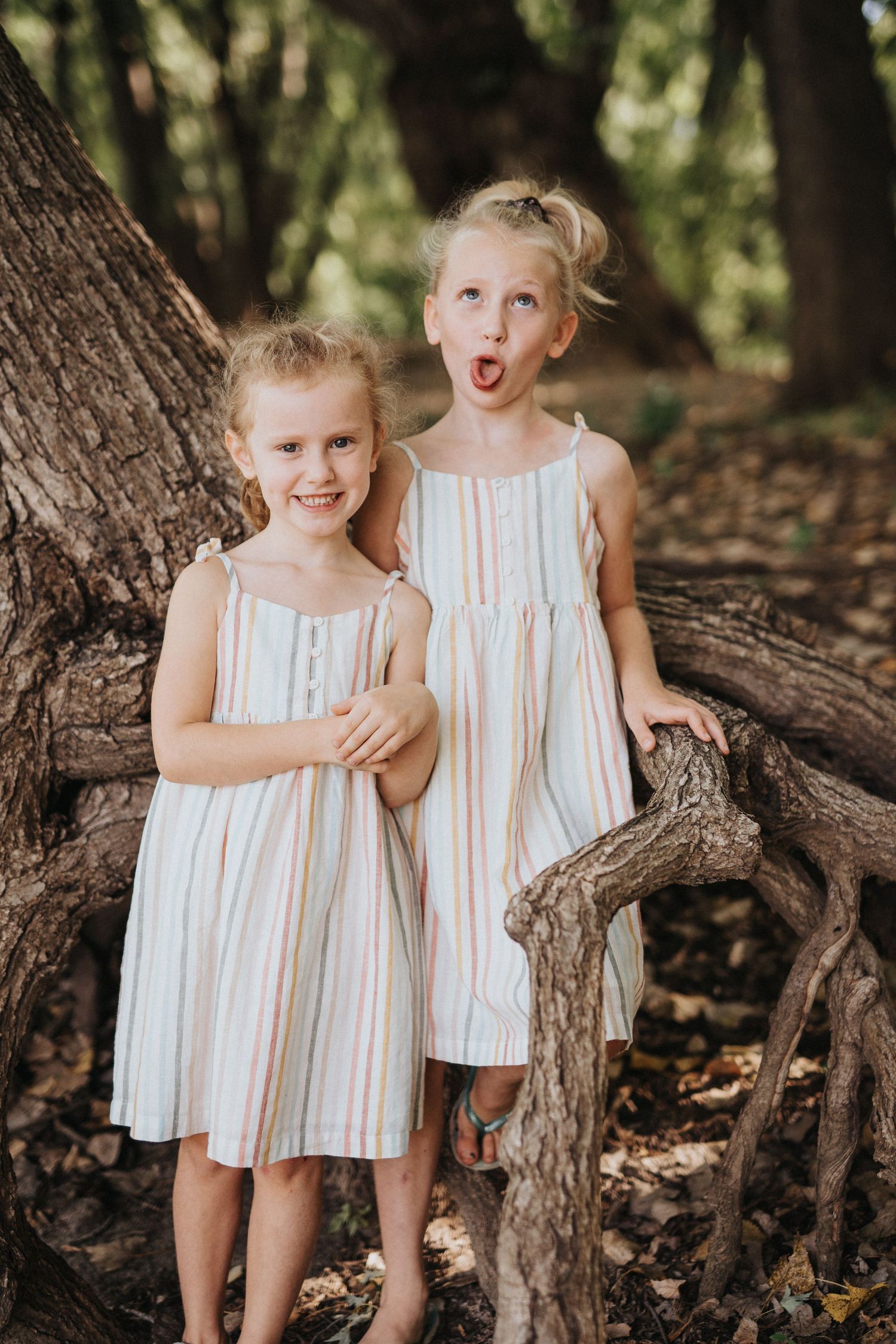 Two children wearing matching white striped summer dresses pose playfully by a large tree trunk in a forest setting.