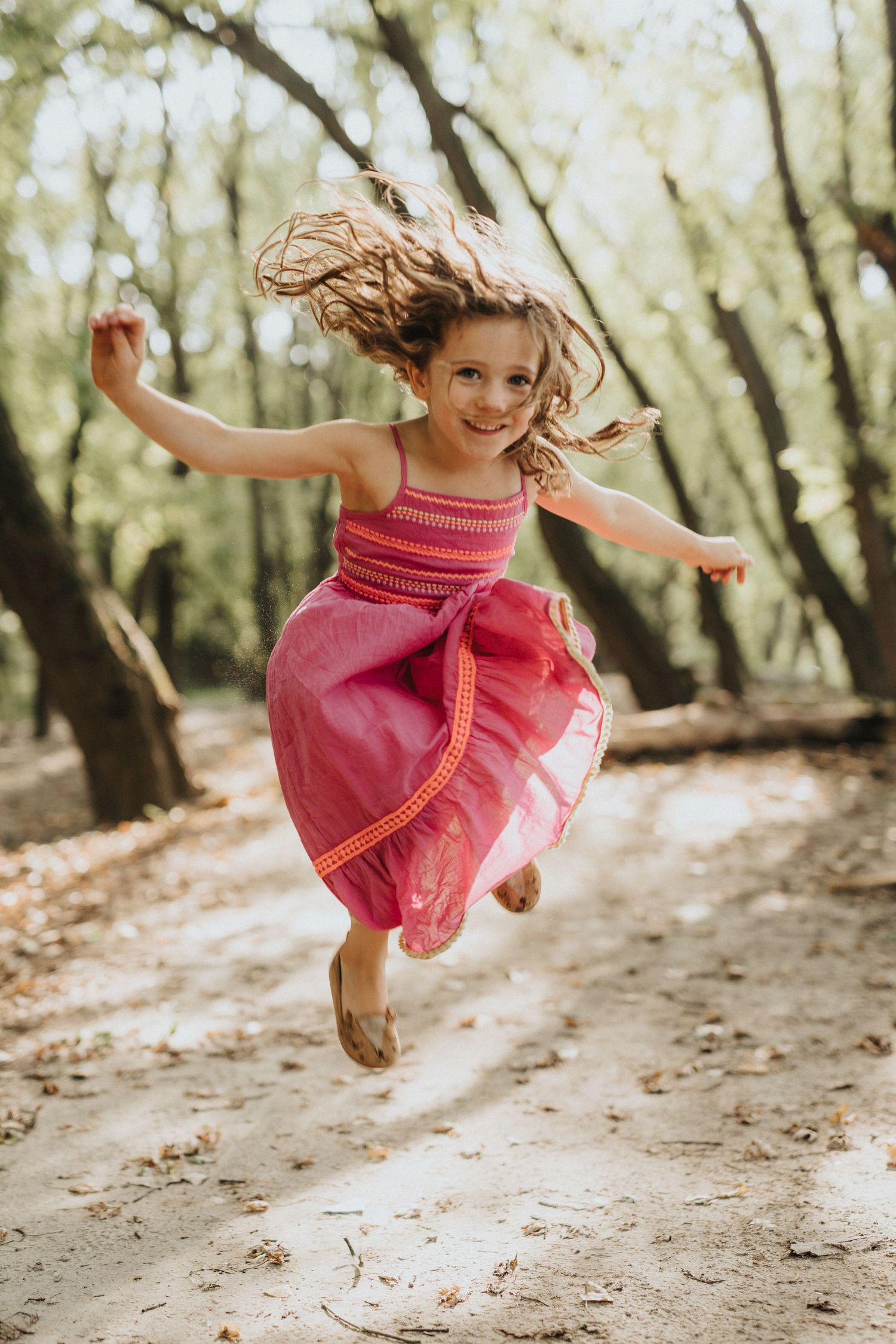 A joyful leap captured in a pink dress on a sunlit forest path, hair flowing in mid-air motion.