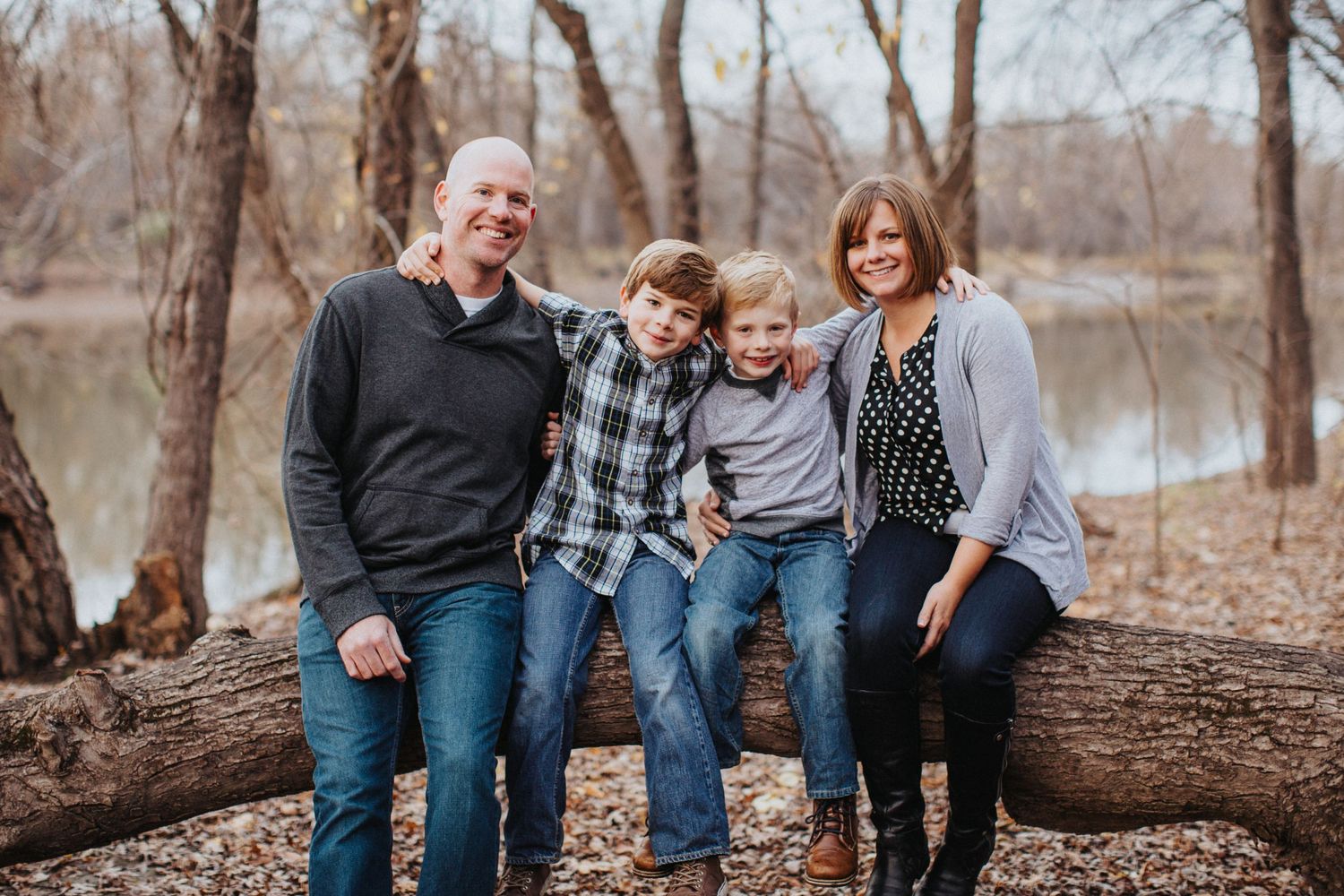 A group of four family members sit together smiling on a fallen tree in a wooded area near a lake on an autumn day.