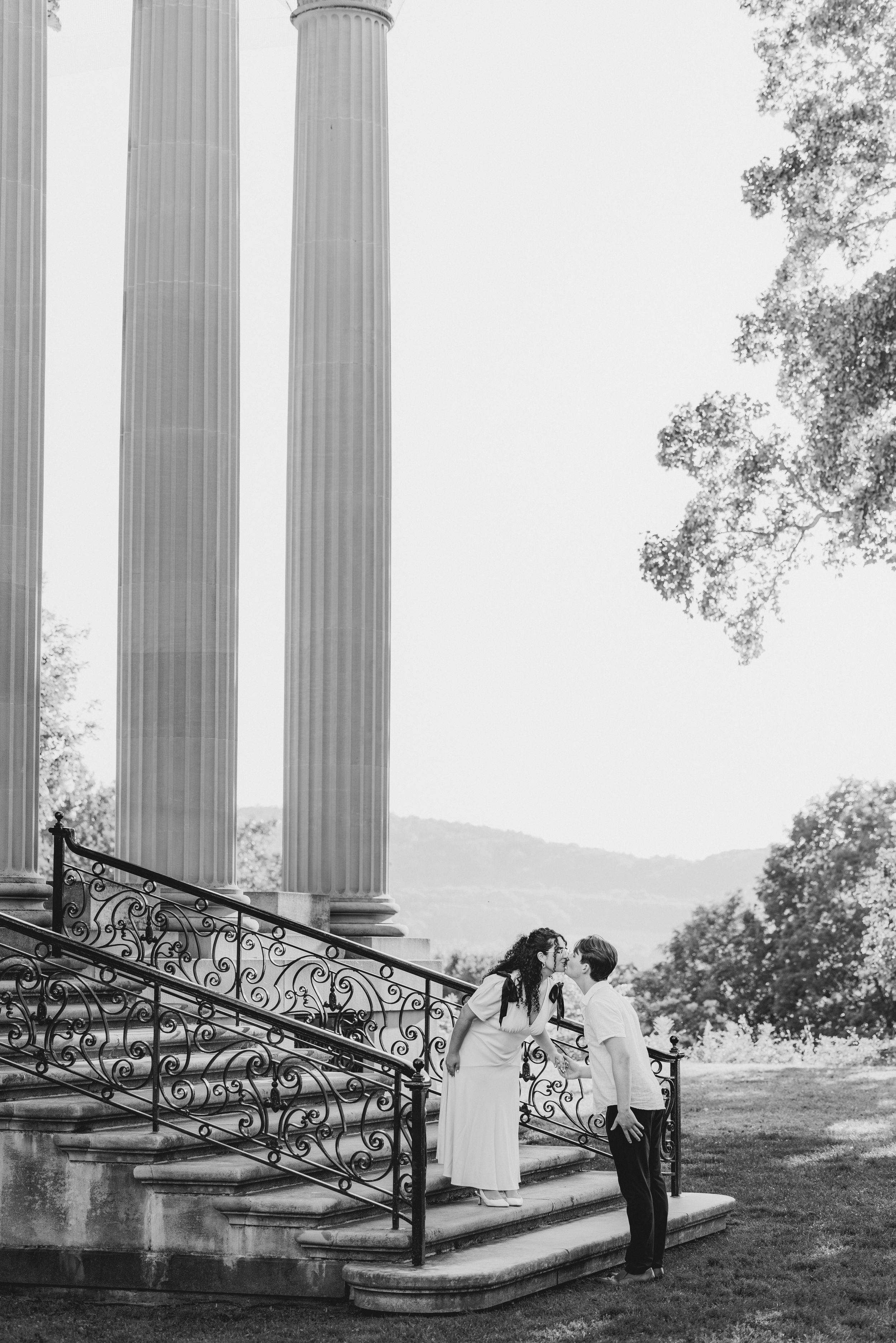 A romantic black and white photo of a couple sharing a tender moment on grand stone steps with classical columns.