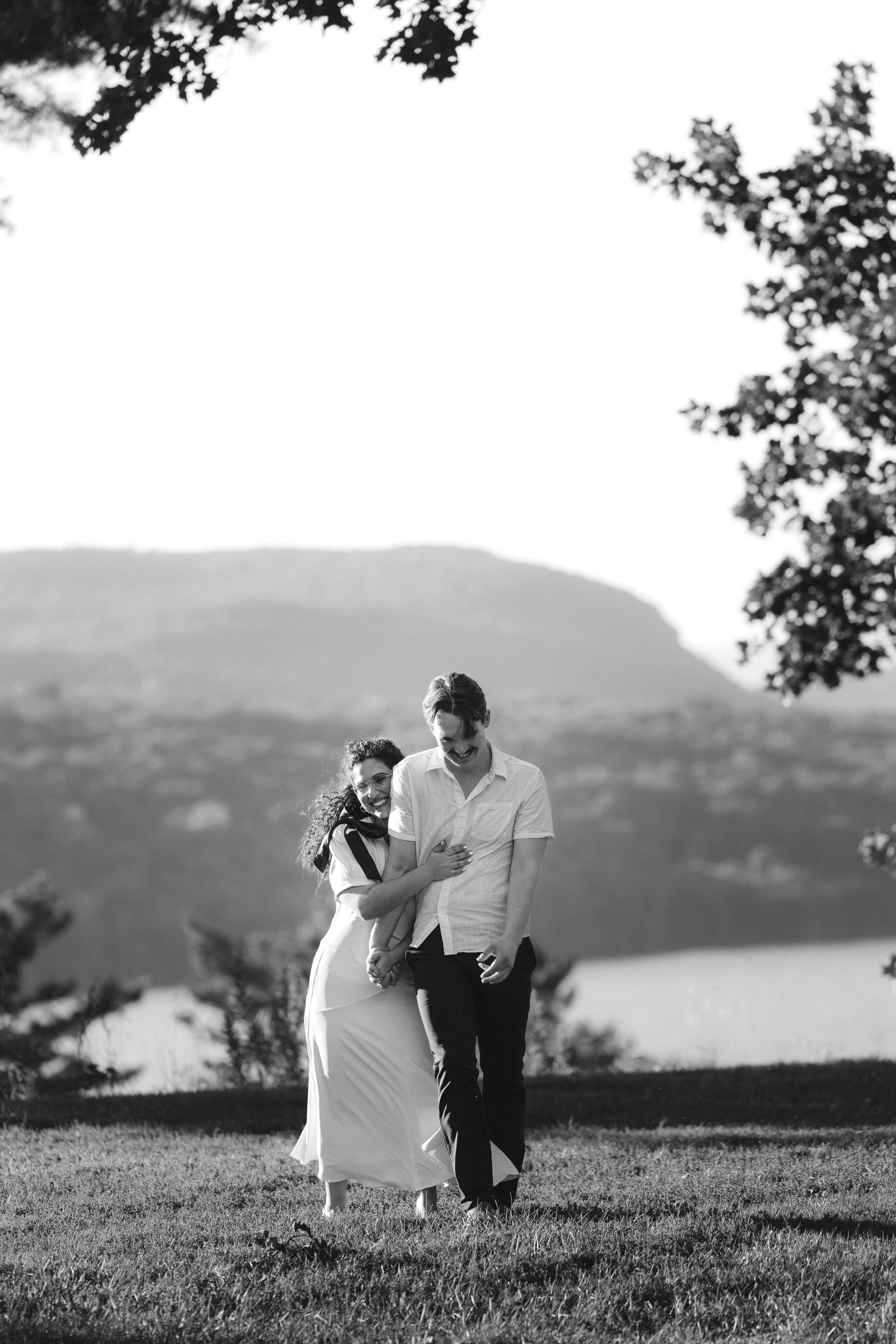 A couple sits together on a grassy hilltop overlooking a scenic mountain landscape in black and white.