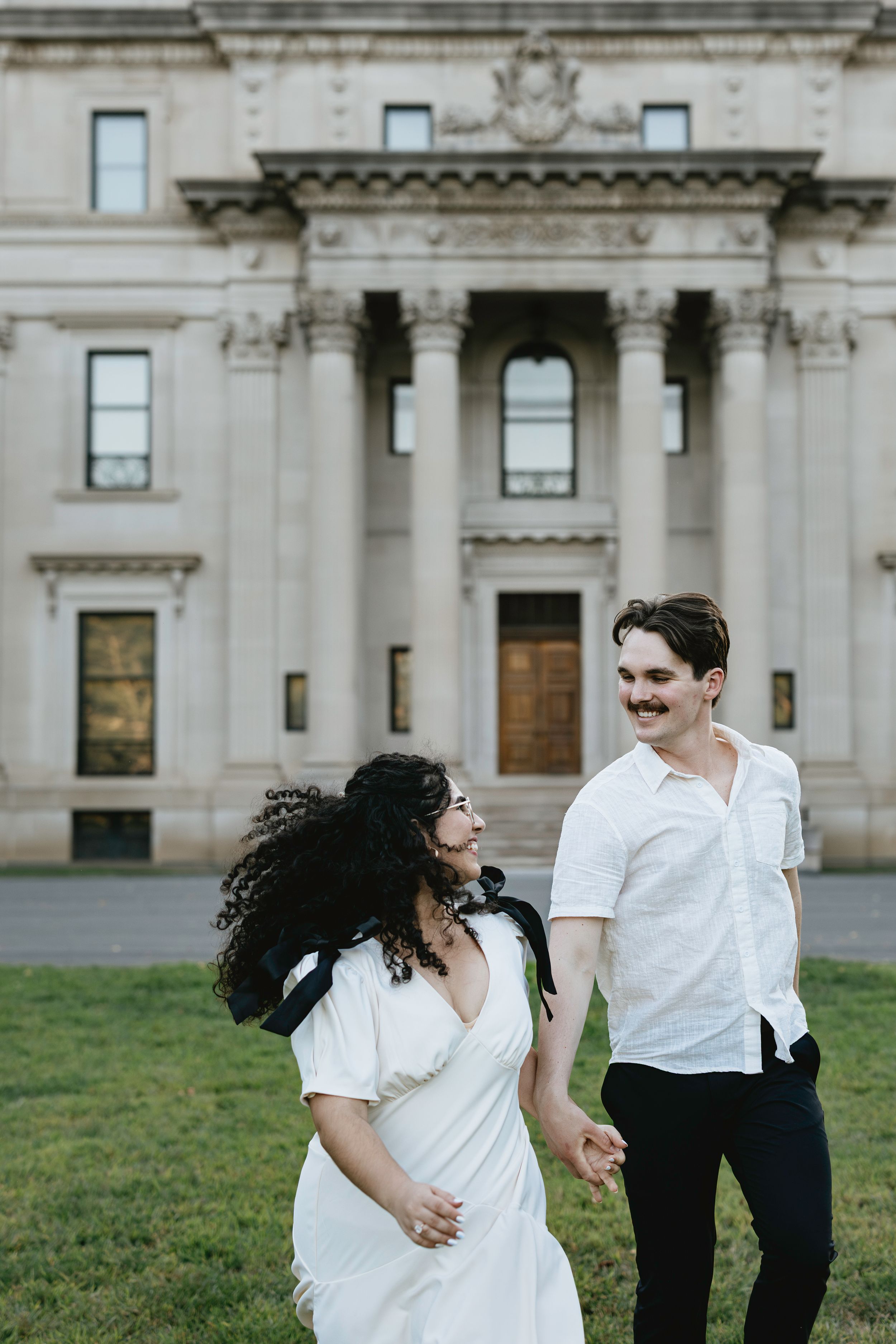 A couple in matching white outfits share a romantic moment in front of a grand neoclassical building.
