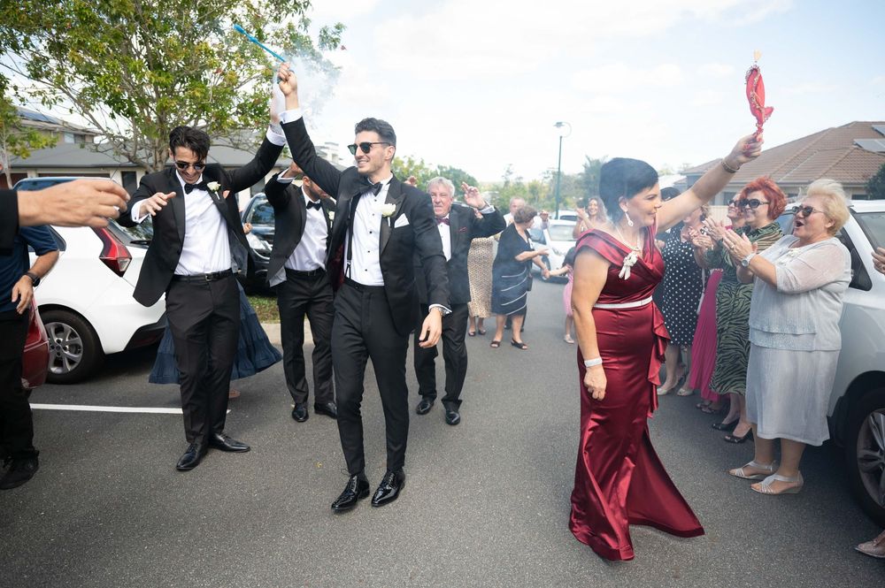 Groom and groomsmen in tuxedos celebrating outside with raised arms at wedding arrival.