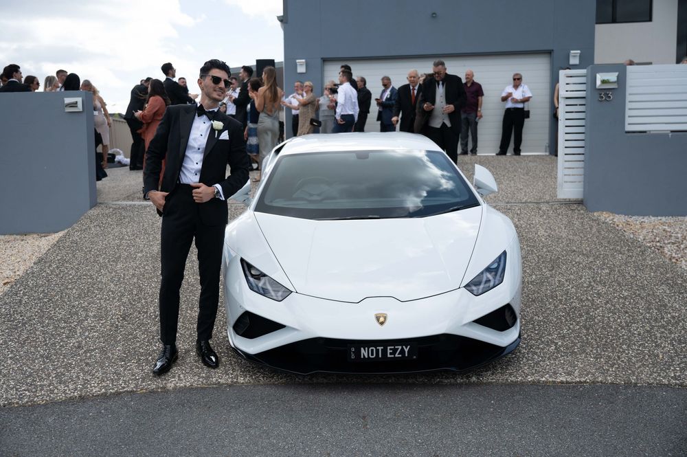 Groom in tuxedo posing proudly beside white Lamborghini Huracan at wedding venue.
