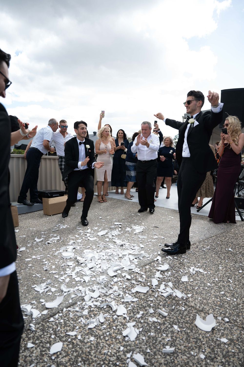Groom dancing joyfully amid broken plates in traditional Greek wedding celebration outdoors.