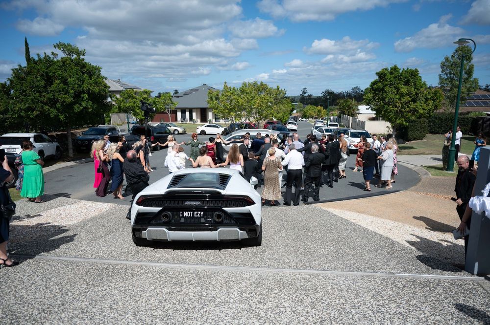 Wedding guests gathered around white Lamborghini Huracan in suburban street celebration.