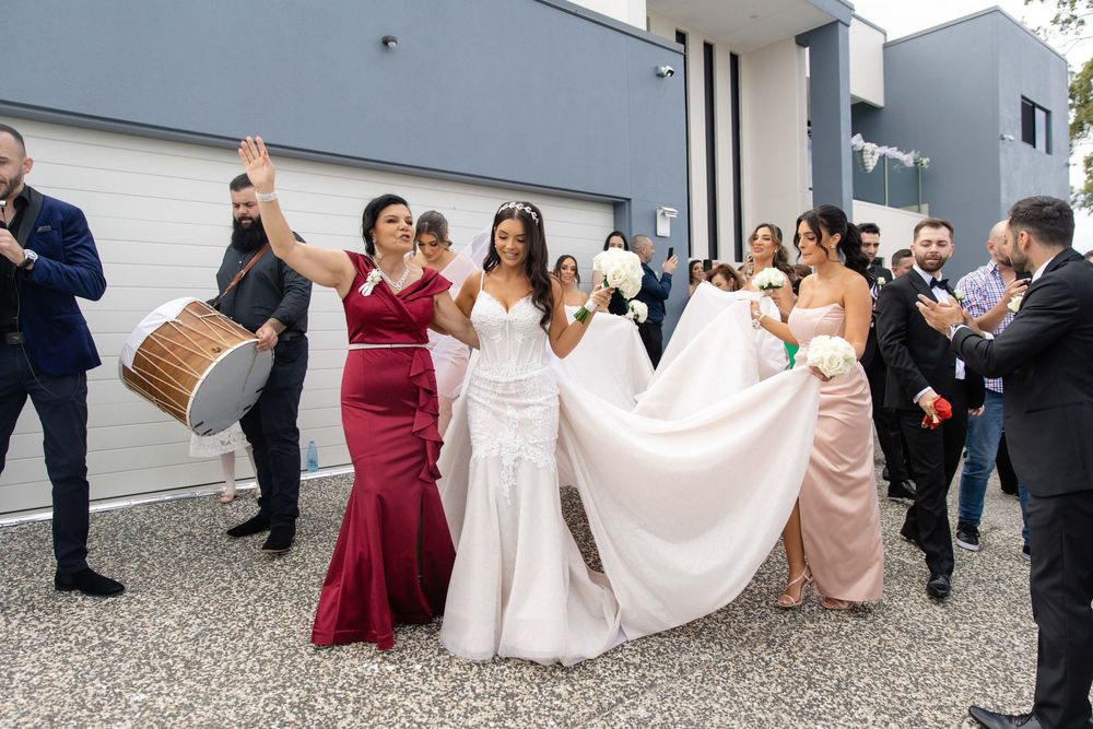Bride in white gown escorted by bridesmaids with traditional drum music outside home.