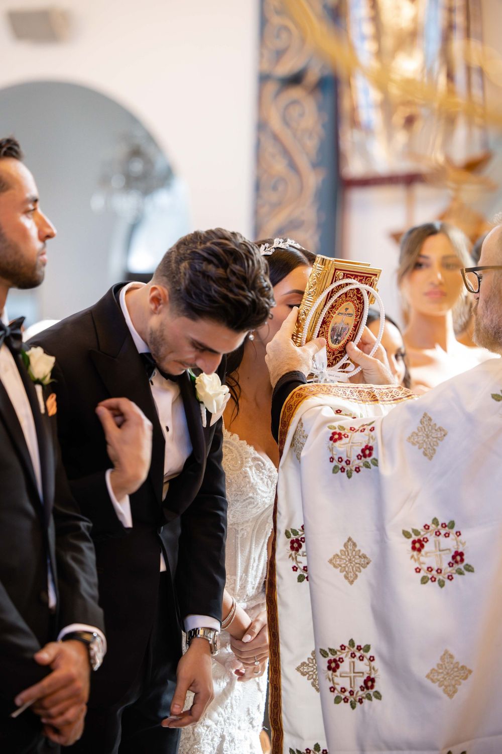 Bride and groom kiss holy book held by Orthodox priest during ceremony.
