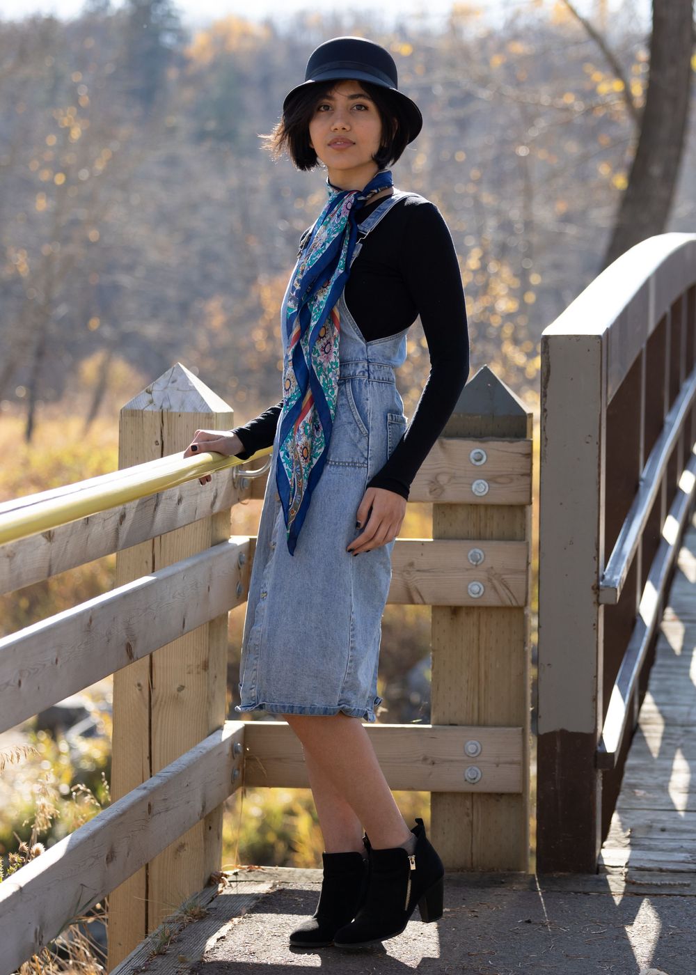 Fashion model in denim vest, black hat and dress poses against wooden railing in autumn outdoor setting.