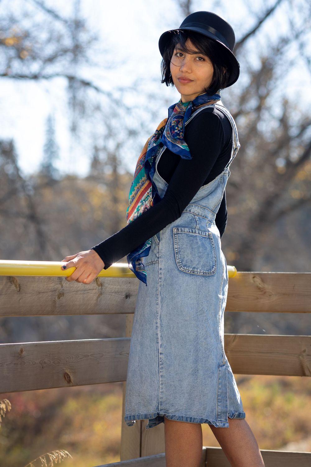 Person wearing denim overalls and black hat poses against wooden railing in autumn outdoor setting.