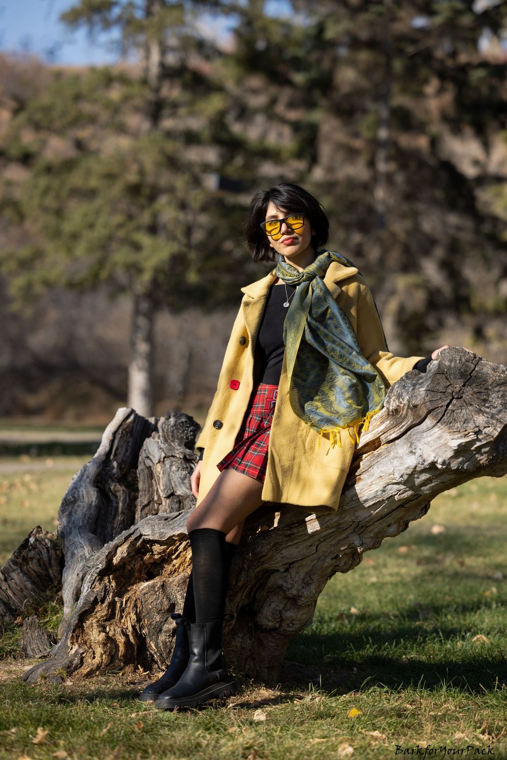 A person in a yellow jacket and boots sits casually on a large fallen tree trunk in a sunny park setting.