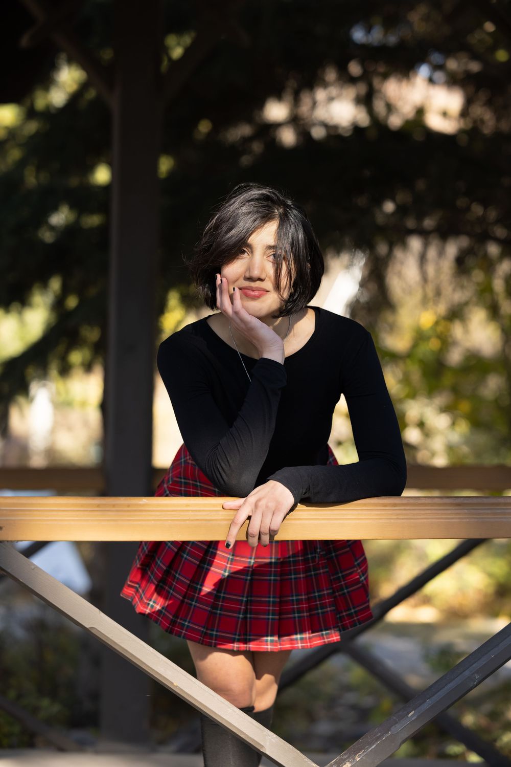 A person in a black top and red plaid skirt leans over a wooden railing in a natural outdoor setting.