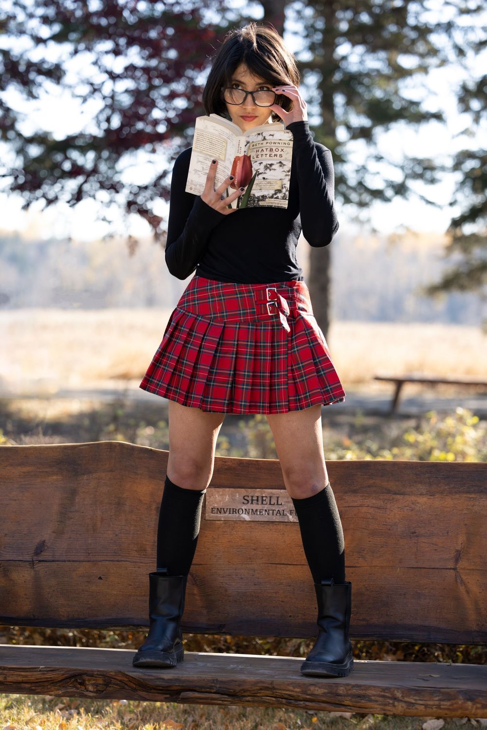 A person wearing a red plaid skirt, black boots and knee socks poses on a wooden bench in an outdoor setting.
