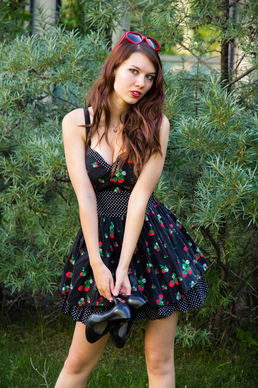 A vintage-style cherry print dress and red sunglasses are worn in a garden setting with green foliage.