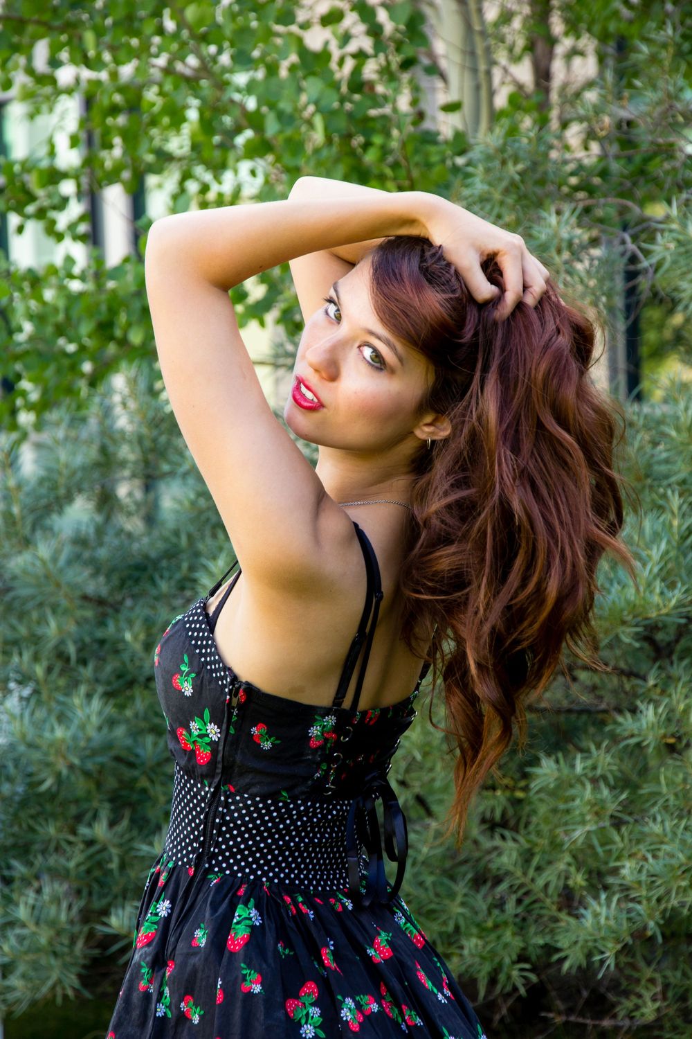 A woman in a black floral dress with polka dots poses outdoors against a green foliage background.