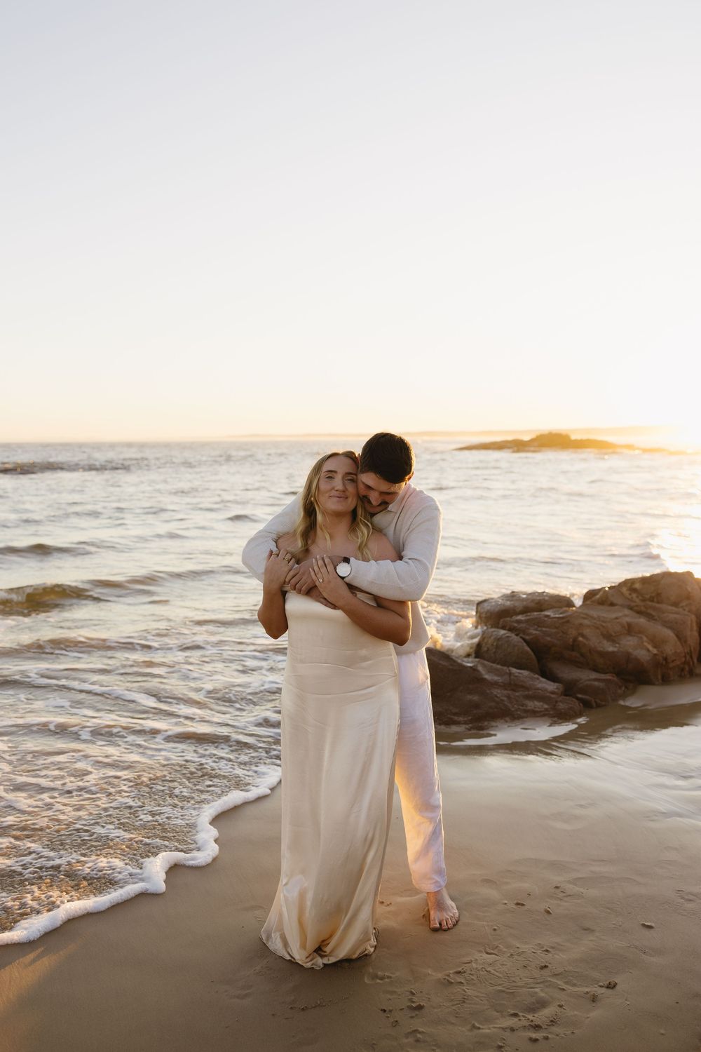 Newlywed couple embracing on a beach at sunset with waves and rocks in the background.