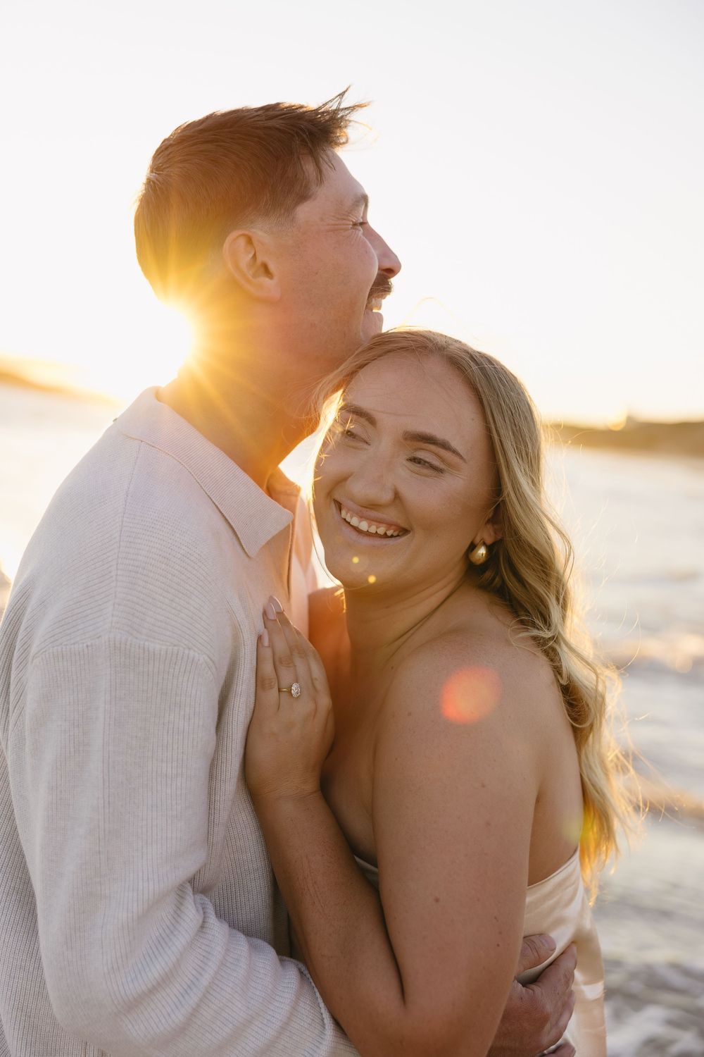 A couple embraces and laughs together on a beach during golden hour sunset with sun flares in the background.